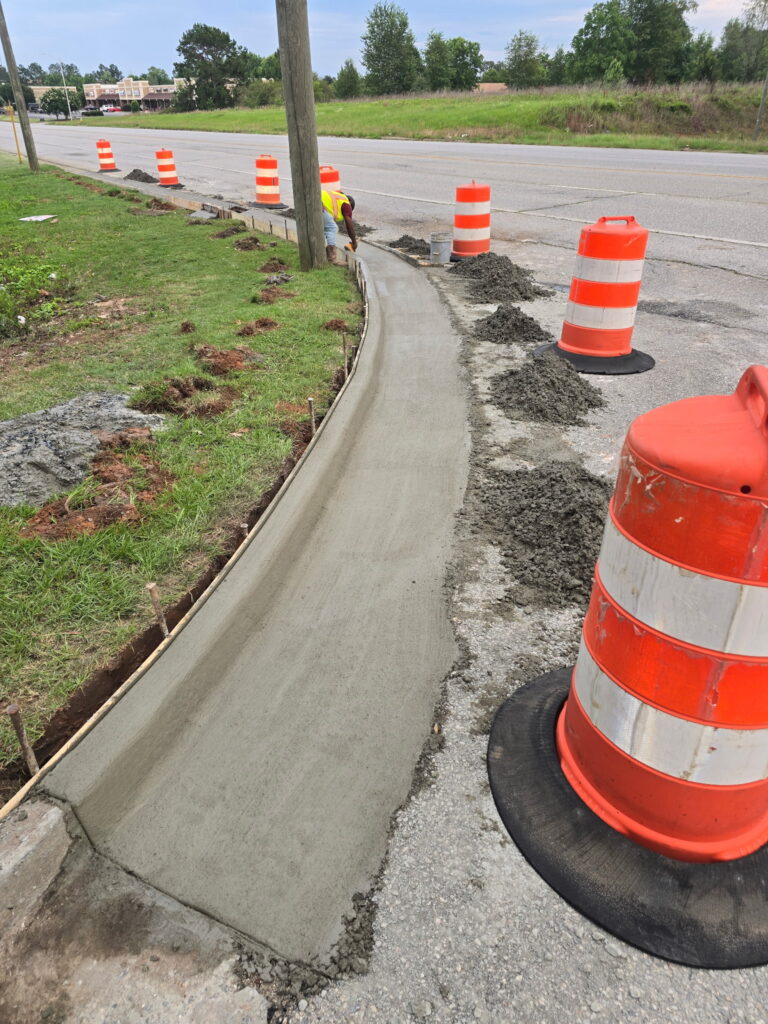 New concrete sidewalk curving along a highway shoulder behind traffic drums